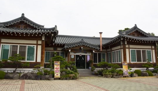 기와지붕을 얹은 한옥 식당 정면 — 가운데 현관과 돌계단, 앞마당의 소나무 분재와 화분